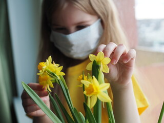 Little girl in medical mask sitting on the windowsill with yellow daffodil flowers. Covid 2021. The concept of pediatric healthcare. Stay at home-quarantine prevention of the coronavirus. Hello spring