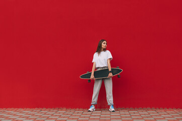 Horizontal full-length portrait of a stylish female skater holding her skateboard and standing on a city street in front of a red wall with a copy space. - Powered by Adobe