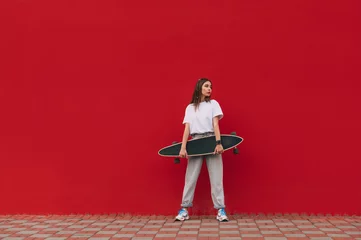 Tischdecke Horizontal full-length portrait of a stylish female skater holding her skateboard and standing on a city street in front of a red wall with a copy space. © bodnarphoto