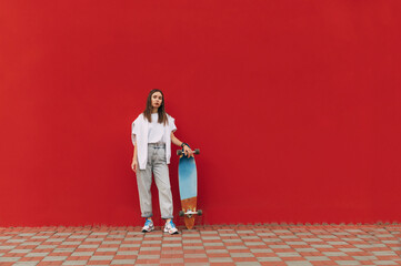 Full-length photo of a stylish skater woman in casual attire standing on a red background, holding her longboard.
