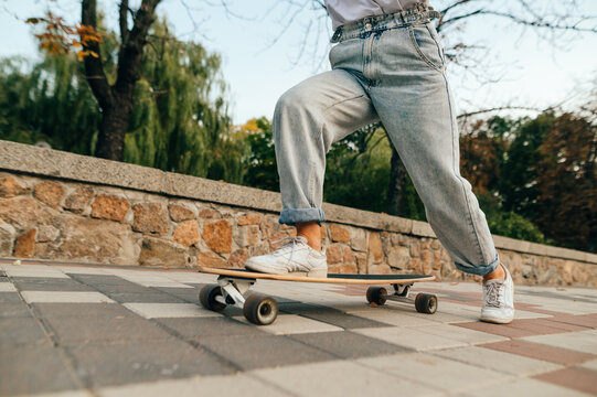 Cropped photo of female skateboarder legs in baggy jeans, riding her long board on a city street near the park.