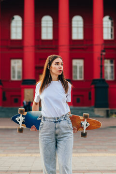 Vertical Shot Of A Skater Woman In Baggy Outfit Holding Her Colorful Longboard, Standing In Front Of A Red Building On A City Street.