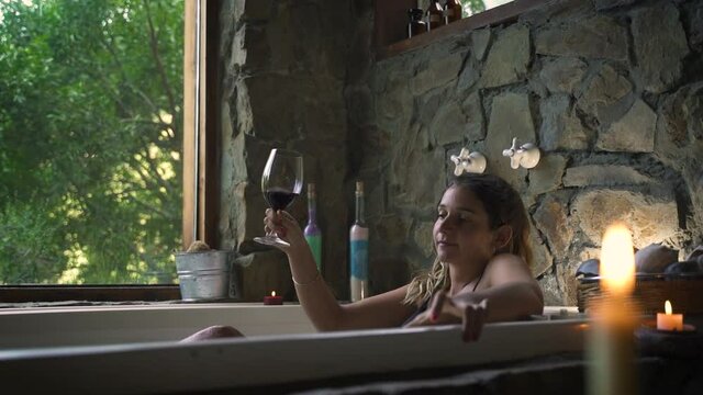 Young attractive female having a fancy bath in a jacuzzi drinking wine with big green nature window able to slow motion 60fps