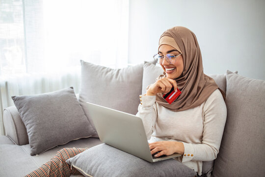 Online Shopping. Smiling muslim woman in hijab using smartphone, laptop and credit card at home, purchasing goods in internet, free space. Arab woman making online purchase on laptop