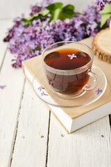 Romantic background with a cup of tea, lilac flowers and a book over a white wooden table. Leisure concept, spring breakfast