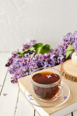 Romantic background with a cup of tea, lilac flowers and a book over a white wooden table. Leisure concept, spring breakfast