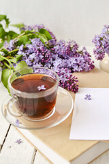 Obraz na płótnie Canvas Romantic background with a cup of tea, lilac flowers and a book over a white wooden table. Leisure concept, spring breakfast. Mockup.