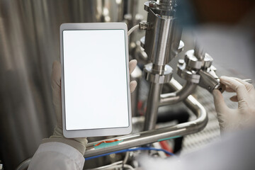 Close up of unrecognizable worker holding tablet with blank white screen while operating equipment at chemical plant, copy space mockup