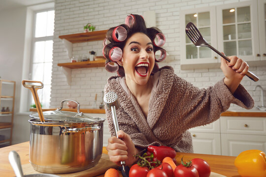 Happy crazy goofy young woman having fun while making healthy vegetable soup at home. Funny positive excited housewife in hair rollers and bathrobe standing at kitchen table, cooking meal and laughing