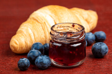 A butter croissant with a personal size jar of blueberry jam surrounded by fresh blueberries