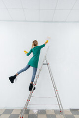A young girl on a stepladder paints a white wall with a roller. Repair of the interior.