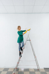 A young girl on a stepladder paints a white wall with a roller. Repair of the interior.