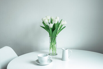 Bouquet of white tulips in a glass vase, a cup and a milk jug on a white table