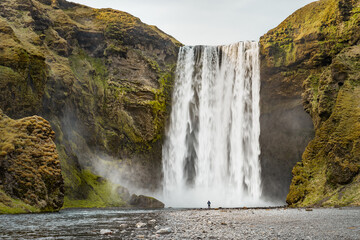 Obraz premium View of famous Skógafoss waterfall with a man standing at the waterfall alone on a sunny spring day. Popular Travel destinations. Classic Icelandic Landscape
