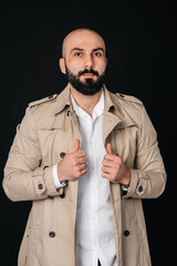 A young bearded man stands and adjusts his raincoat against a black background.