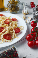 Delicious tagliatelle pasta with cherry tomatoes and basil on a white plate on a wooden table along with pasta ingredients: olives, tomatoes, garlic, olive oil and different types of dried peppers.