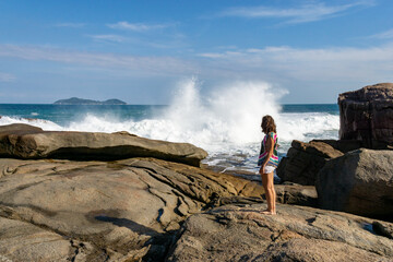 Mujer con pantalones cortos blancos y camisa colorida mira romper una ola en las rocas de la costa