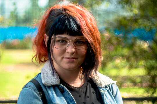 Portrait of a young caucasian woman with black and red hairstyle, jean jacket and vintage glasses looking at camera with serious expression. Green trees and park in the background