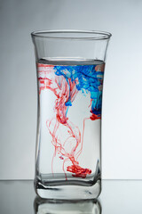 Single drops of light blue and red ink falling down into the glass full of water in the studio. Reflecting surface and gradient background