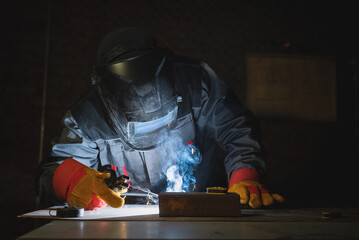 Welder is welding a metal pipes on the workbench in the dark.