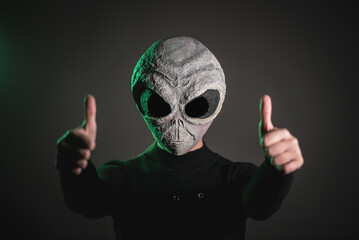 An alien is showing a thumbs up gesture on the dark background.