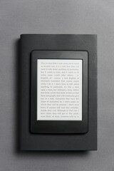 Electronic reader over a closed black traditional book, on a gray background.