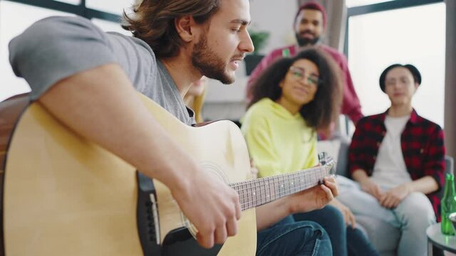 Close up of handsome boy plays the guitar and sings songs. Friends have fun and relax at home. Multiracial friends cheerfully sit at home and sing songs to the guitar