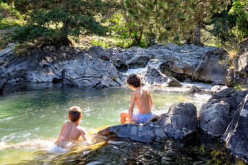 two boys on their backs enjoying a swim in a clear mountain river in a rocky, sunlit pool with rocks all around