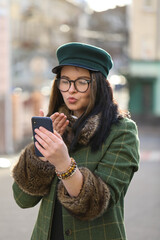 Beautiful woman in coat and hat uses smartphone while walking in spring city. She takes a selfie or makes a video call .