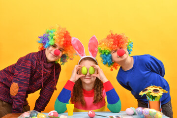 Girl holding Easter eggs in front of her eyes two clown boys are standing side by side.