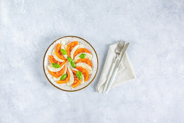 Caprese salad with sliced fresh tomatoes, mozzarella cheese and basil served on a white plate on light gray table surface, top view.