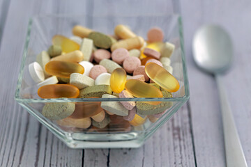 Assorted colorful pharmaceutical medicine pills, tablets and capsules in glass bowl whith spoon on the table; color photo.