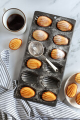 Flat lay with madeleine cakes in a baking tin, coffee a plate of cakes and a tea towel
