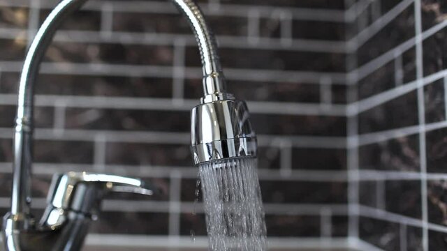 Close-up of tap water flows to sink on blurred background of kitchen tile wall.