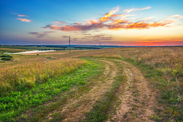 Rural landscape with ground road and wheat field in a countryside at summer sunset