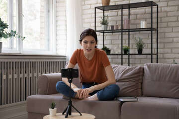 Video blogger. Inspired young woman sit on sofa cross legged talk speak in front of cellphone web...