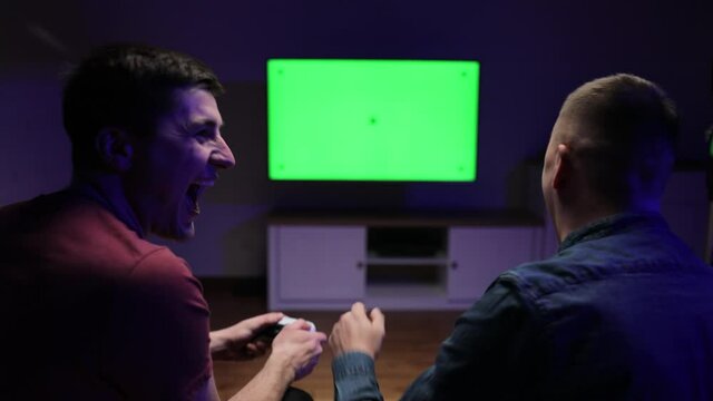 Happy young people playing video games on console while sitting on couch in front of tv. On Video Game Championship Professional Gamer Plays on Green Chroma Key Screen Personal TV
