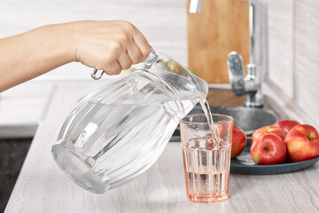 A woman's hand pours drinking water from a clear glass decanter into a glass in the kitchen of her home. In the background is a plate of fruit. The concept of a housewife. Selective Focus