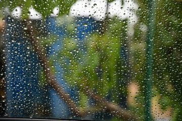 Blurry view through transparent glass window surface covered with shiny raindrops from behind. Defocus texture of water drops with blurred blue green background with copy space
