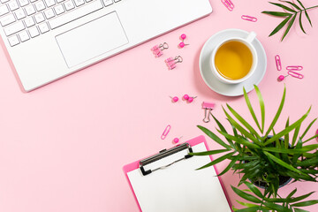 Creative office desktop with stationery set, clipboard with blank paper sheet, green potted plant and cup of aromatic tea on pink pastel background.