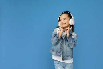 cute smiling little child girl in white headphones and denim listening to music and dancing on blue background. copy space