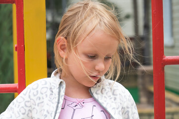 a little girl with blond hair, 5 years old, on the street, against the background of a playground, lowered her head and looks down