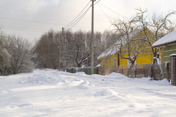 A cloudy winter day with snowfall. A street in a dacha village. Snow covered rural road and snowdrifts along the road near the fences.