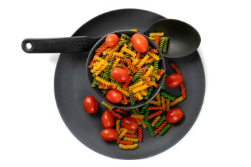 overhead view of a black bowl of raw colored fusilli pasta and small tomatoes isolated on white

