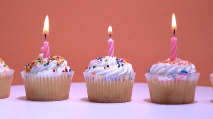 Birthday cupcakes with burning candles on orange background. Vanilla coconut muffins with white whipped cream and sugar sprinkles icing. Close-up shot with shallow depth of field.