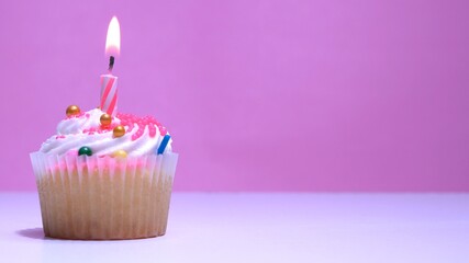 Cupcake with burning festive candle at white cream and sugar icing on pink background, macro shallow depth of field view. Homemade vanilla cup cake with buttercream frosting close up.