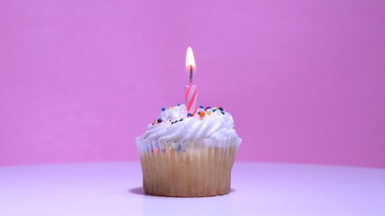 Cupcake with burning festive candle at white cream and sugar icing on pink background, macro...