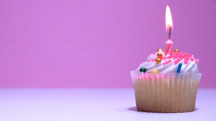Cupcake with burning festive candle at white cream and sugar icing on pink background, macro...