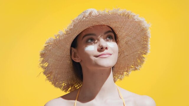 Young good-looking brunette caucasian woman in a straw hat puts spf cream on her cheeks, enjoys the sun and smiles for the camera against yellow background  | Spf cream concept