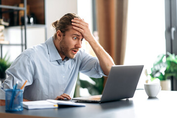 Shocked annoyed caucasian busy guy looking at a laptop in confusion, received unexpected news, failed a deal, shouts, holds his head with his hand, sitting at the work desk